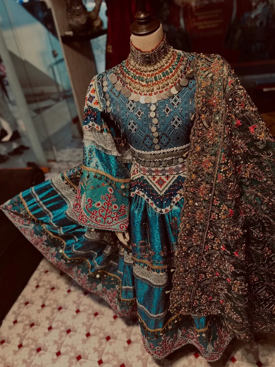 Hand-embroidered traditional Afghan women's dress with coin jewelry, displayed on a mannequin in a Kabul shop