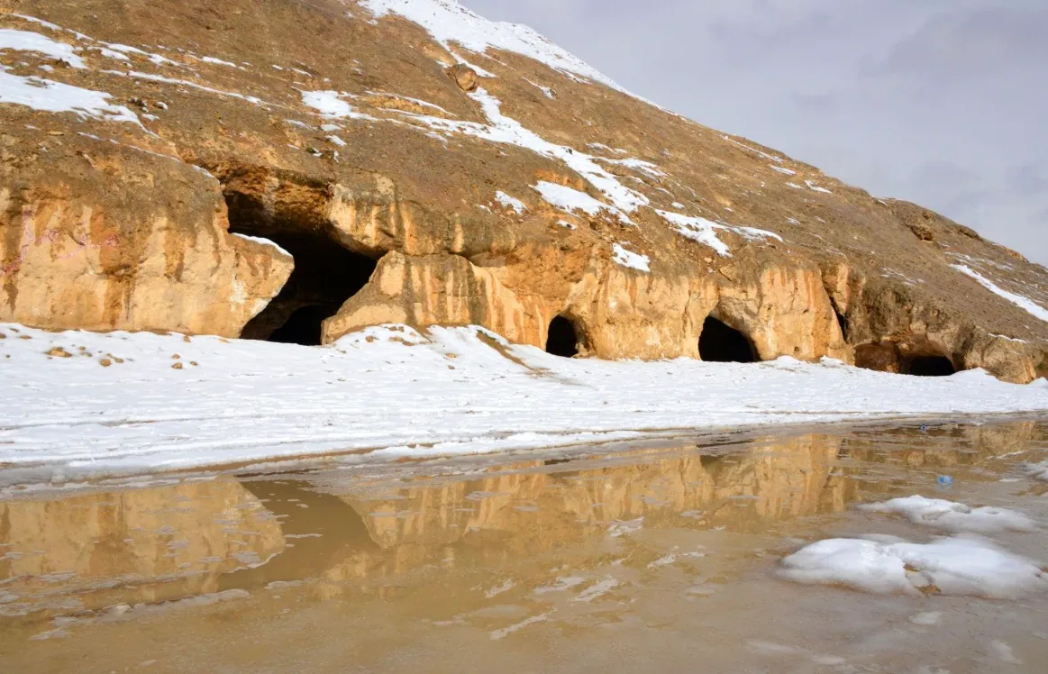 Cave openings cut into a hillside in Samangan, with snow on the ground reflected in meltwater