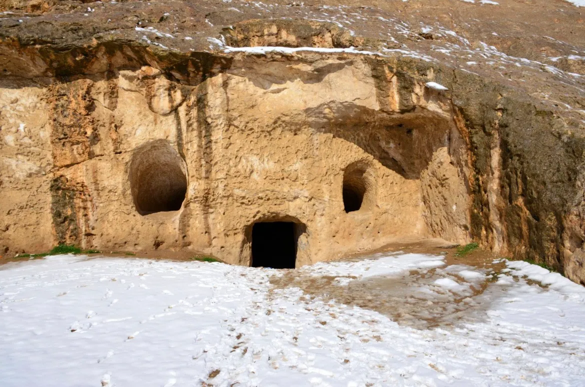 Cave doorways cut into a rock face, with snow on the ground below, in Samangan